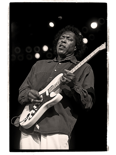 Black and white image of blues musician Buddy Guy passionately playing an electric guitar on stage. He is wearing a long-sleeved shirt and has eyes closed, focusing intensely on the performance.