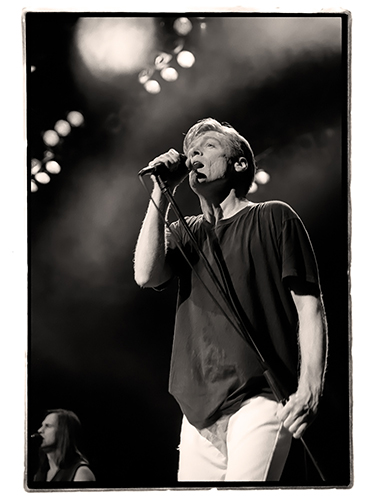 Black and white photo of Bryan Adams wearing a dark shirt and light pants, passionately performing on stage with a microphone in hand. Stage lights shine down, creating a dramatic atmosphere.