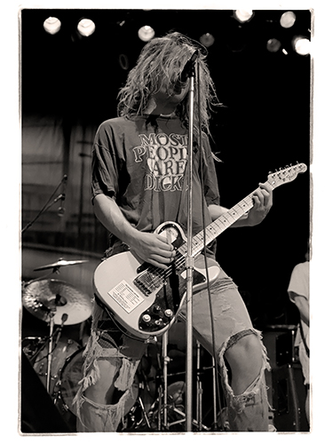 Black and white photo of Dave Pinner performing on stage. He has long hair, wear a shirt with text, and play an electric guitar. The stage includes a drum set in the background.