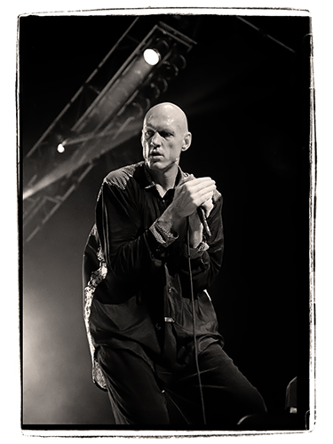 Black and white photo of Peter Garrett passionately into a microphone on stage. He wears a dark, loose-fitting outfit, and the lighting casts a dramatic shadow. 