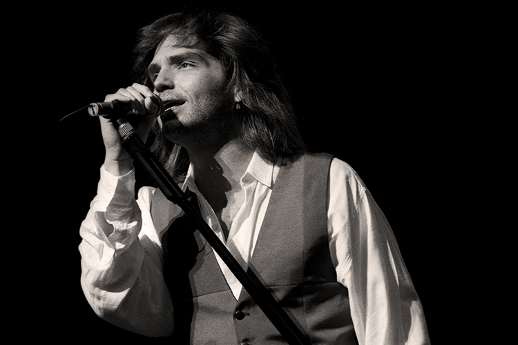 A black-and-white image of Richard Marx performing on stage, holding a microphone stand. He is wearing a vest over a collared shirt, captured against a dark background.