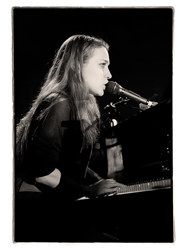 Black and white image of a Fiona Apple playing piano and singing into a microphone on stage.