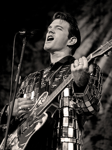 Black and white photo of Chris Isaak passionately singing into a microphone while playing a guitar. He is wearing a jacket made of shiny, rectangular mirrors, and the background is softly blurred.