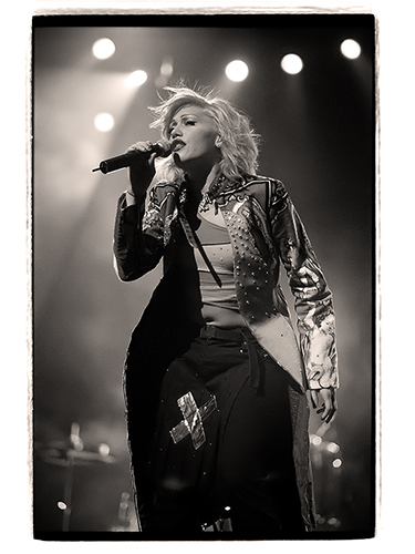 Black and white photo of Gwenn Stefani performing on stage. She wears a long, patterned coat and holds a microphone, with stage lights illuminating the background