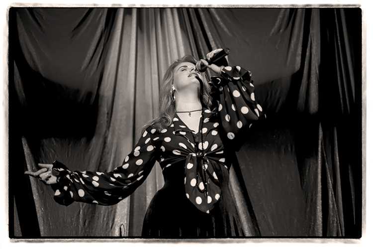 Trisha Yearwood singing passionately on stage, holding a microphone. She is wearing a polka dot blouse and earrings, with draped curtains in the background, captured in black and white.