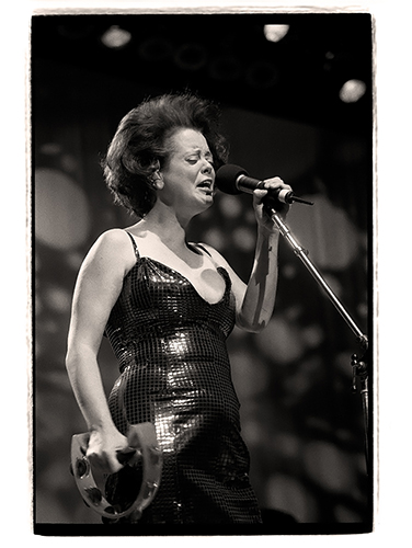 Cindy Wilson in a sequined dress sings passionately into a microphone on stage while holding a tambourine. The black-and-white image captures her mid-performance.