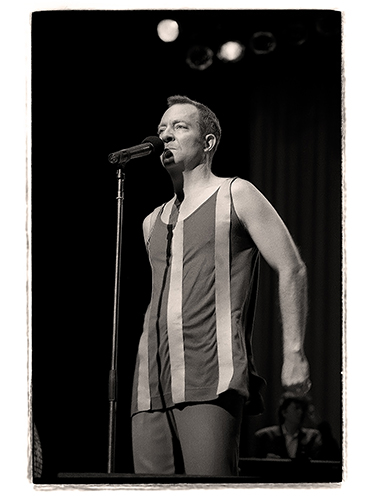 Fred Schneider performing on stage. He is wearing a sleeveless outfit with vertical stripes. The image is in black and white, with a blurred background and stage lights visible above.