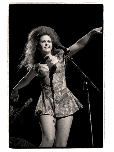 Performer Katie Pierson dancing freely next to a microphone on stage, wearing a patterned dress, fishnet stockings and voluminous hair. The black and white image captures a dynamic pose with one arm extended