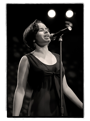 Natalie Merchant stands on stage singing into a microphone. She is wearing a sleeveless dress and appears focused. The background is dark with bright stage lights