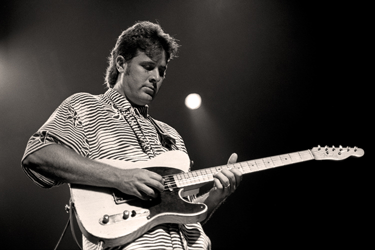 A black and white image of a Vince Gill playing an electric guitar on stage. He is wearing a striped shirt and focused on the performance, with a spotlight shining above him against a dark background