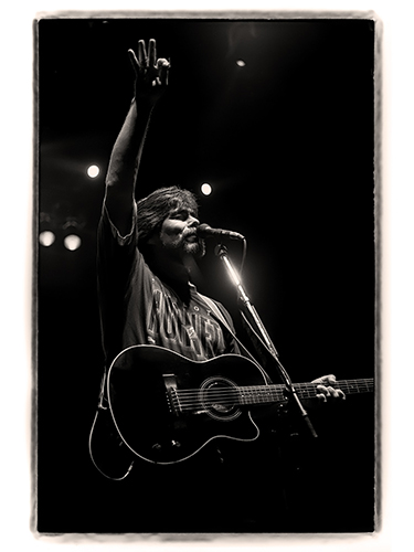 Musician Randy Owen plays an acoustic guitar on stage. He is singing into a microphone and raising one hand with three fingers extended. The lighting is dramatic, highlighting his figure against a dark background.