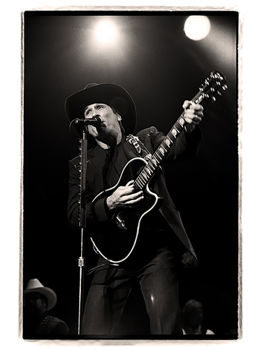 Black and white image of a Clint Black wearing a cowboy hat and playing an acoustic guitar on stage. He is singing into a microphone, with stage lights shining in the background.