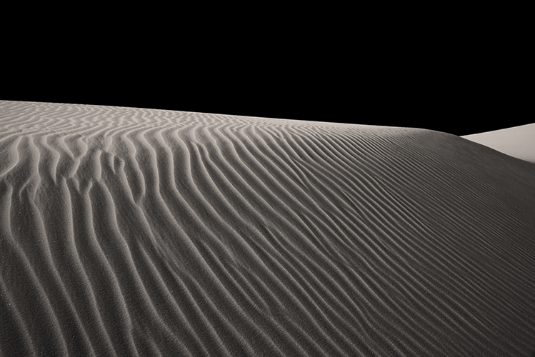 A white sand dune under a dark sky. The foreground shows textured ripples in the sand. 