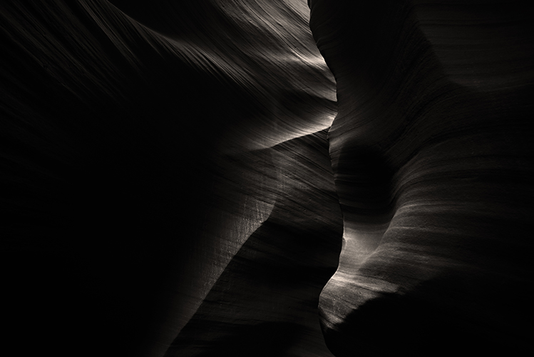 A dramatic and abstract view of a slot canyon interior, highlighting dark, smooth rock surfaces with soft, undulating lines.