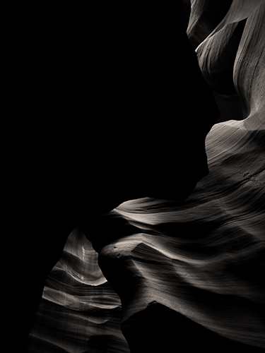 A partially obstructed view of the interior of a slot canyon highlighting the dark, smooth rock surfaces with soft, rolling lines. 