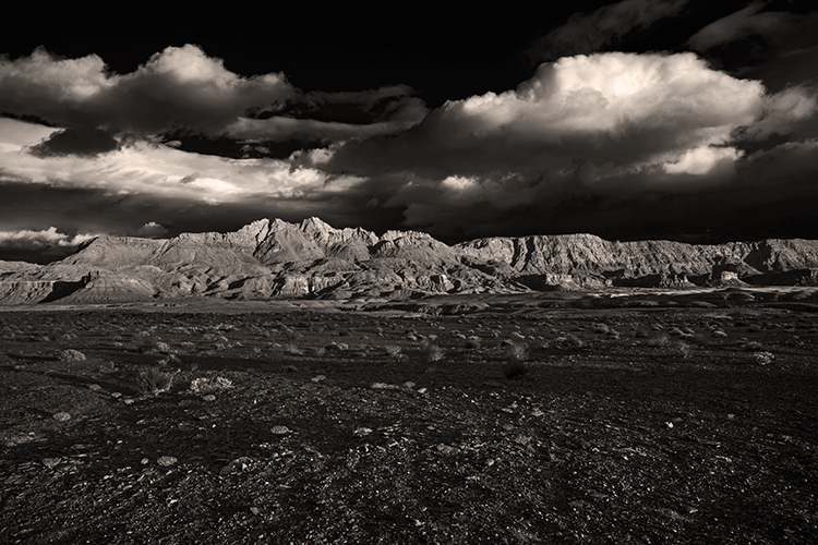 A desert landscape with mountains in the distance under dark skies and white puffy clouds. The light cast long shadows along the foreground with rocks and patches of grass.