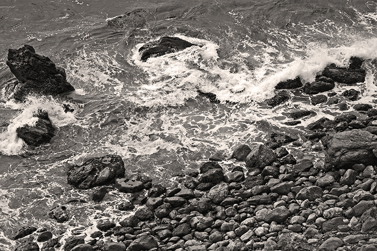 Black and white image of ocean waves crashing against rocky shore. The surf foams over large, dark rocks scattered along the coastline, creating a textured and dynamic scene.