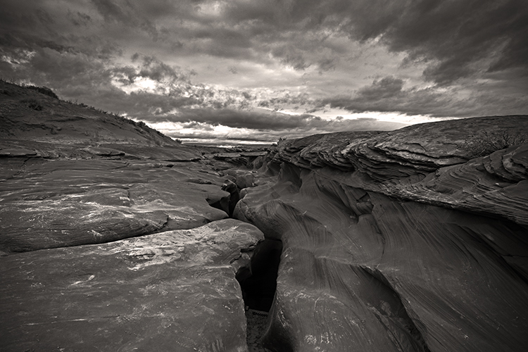 A dramatic black and white landscape photograph depicts rugged rock formations under a moody, cloud-filled sky. The terrain features deep fissures and layered textures, creating a stark and powerful scene.