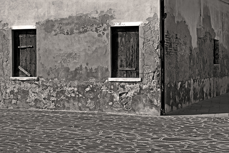 A grayscale image of an old building corner with two shuttered windows. The walls are weathered and cracked, showing signs of age. The ground is paved with stone tiles, adding texture to the scene.