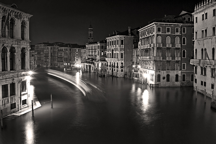 A black and white photo of the Grand canal in Venice at night. Historic buildings line the canal, while streaks of light from a moving boat create dynamic lines on the water, adding contrast to the stillness of the scene.