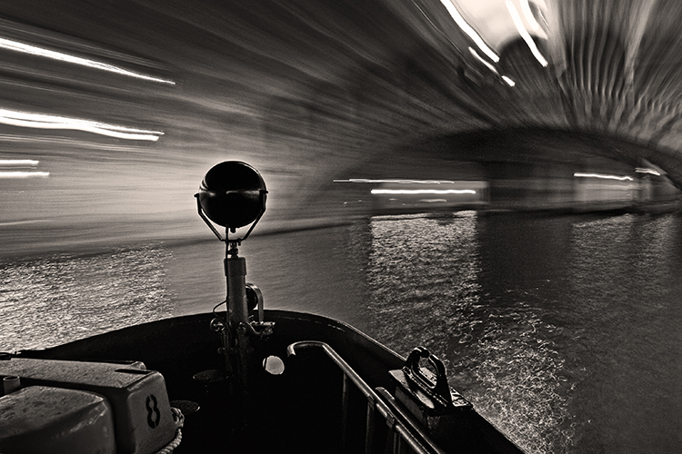 Black and white image of a boat navigating under Rialto Bridge at night. The scene is motion-blurred, capturing the light trails and reflections on the water, creating a dynamic and abstract effect.