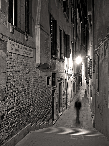 A narrow, dimly lit alleyway at night in Venice, Italy with a blurred silhouette of a person walking towards steps. The walls are lined with shuttered windows creating a moody, mysterious atmosphere.