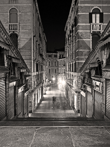 The narrow, deserted walkway atop Rialto Bridge at night lined with tall, historic buildings. The ghost-like shapes of human figures stand at in middle of the street.