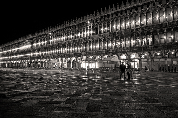 A black-and-white photo of a nighttime scene in St. Marks Square, Venice. The illuminated arches of the Procuratie buildings line the square. Two silhouetted figures stand on the wet, reflective pavement, adding depth to the scene.