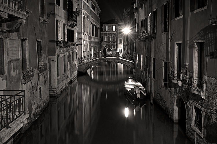 A serene, black-and-white photograph of a narrow Venetian canal at night. A small boat floats on the water, lined by old buildings. A single streetlamp illuminates the scene with a soft glow near a bridge in the background.