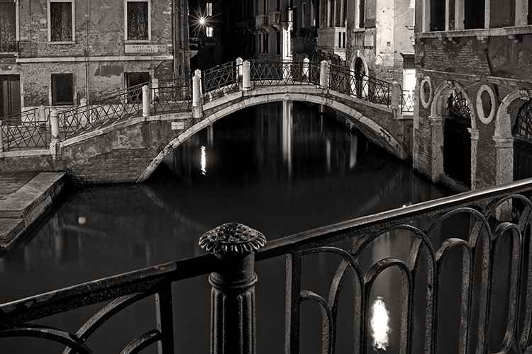 A black-and-white image of a quiet canal at night in Venice, featuring an arched stone bridge with ornate railings. Buildings line the canal, and their reflections shimmer in the water, creating a serene and historic atmosphere.