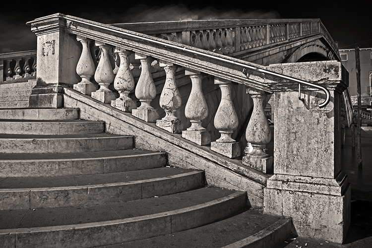 Black and white image of a stone staircase with a carved balustrade, leading up to an arched bridge. The architectural details show ornate, weathered stones and a sturdy handrail. The scene is dramatic with deep shadows and highlights.