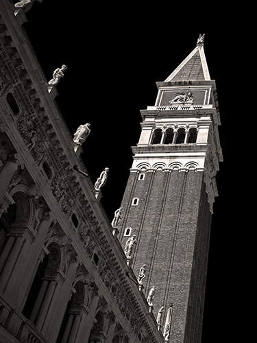 A dramatic black and white photograph of the San Marco belltower in Venice, Italy. The edge of a building featuring intricate carvings and archways with statues along the edge is in the foreground.