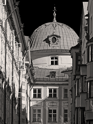 Black and white photo of a historical building with a large, domed roof. The structure is nestled between other ornate, older buildings, highlighted by sunlight.