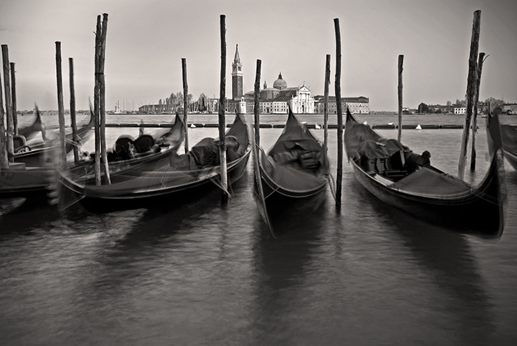 Black and white photo of gondolas moored on calm water in Venice, Italy. The blurred movement of boats creates a serene atmosphere. In the background, the historic church and tower of San Giorgio Maggiore.