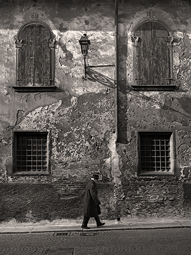 A person in a long coat and hat walks along a cobblestone street in front of a weathered building with two shuttered windows and two barred windows. A vintage street lamp casts a shadow on the textured wall.