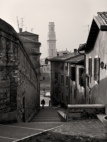 Black and white photo of a narrow, cobblestone street lined with old buildings. A lone figure walks down the stairs towards a distant bell tower. The scene appears to be in a historic European town.