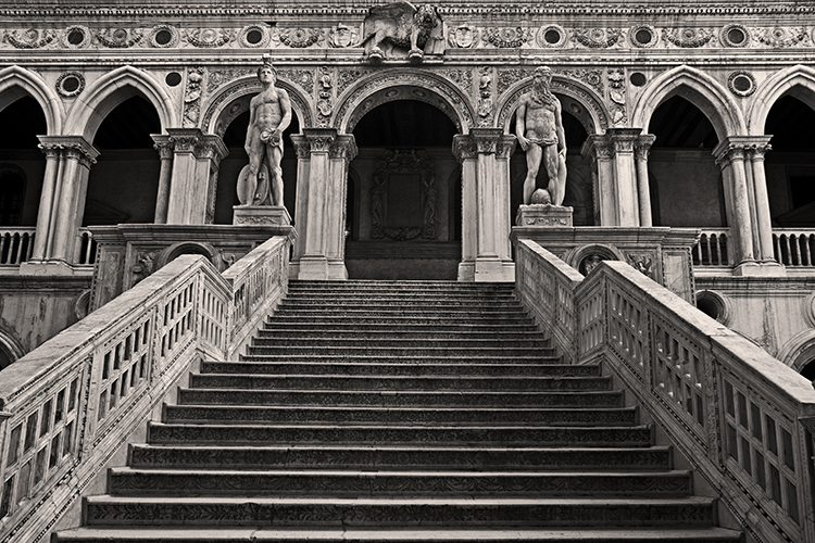 A black and white image of a grand stone staircase leading up to an ornate building facade. Two statues stand on either side at the top of the stairs. The building features intricate carvings and archways.