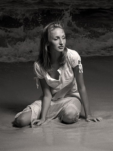 A woman in a white dress sits on the beach, with waves in the background.