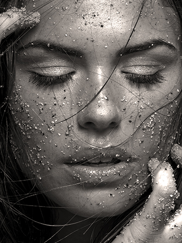 A close-up photo of a woman with eyes closed, covered in sand and water droplets. Long hair strands frame their face. Hands with sand touch their face.