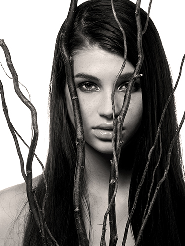 A woman with long dark hair stares through several bare branches, obscuring part of her face. She is gazing attentively at the camera.