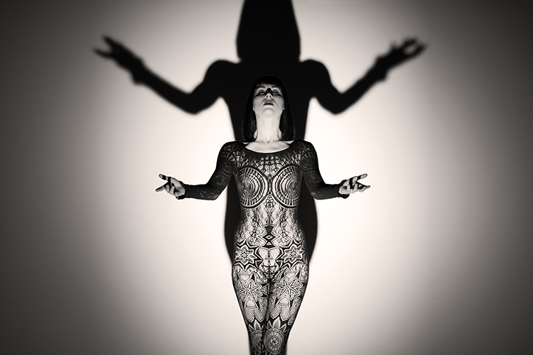 A woman in a patterned bodysuit stands against a wall, casting a dramatic shadow with outstretched arms. The mood is slightly ominous. 