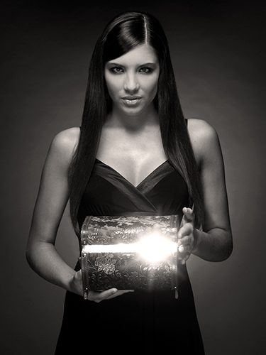 A woman with long, straight hair stands in a dimly lit setting, holding an illuminated decorative box. She wears a dark dress and looks directly at the camera