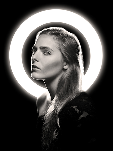 A black and white portrait of a woman with long hair, illuminated by a bright circular light behind her, creating a halo effect. She gazes at the camera with a calm expression.