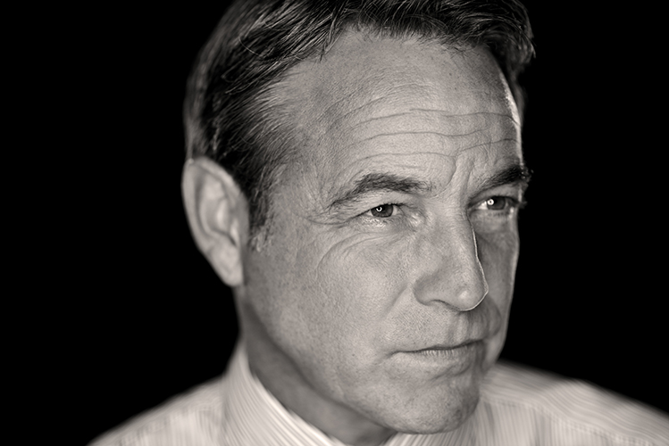 Black and white portrait of a man in a collared shirt, looking to the side with a serious expression. The light and soft focus highlight his eyes and the lines on his face.