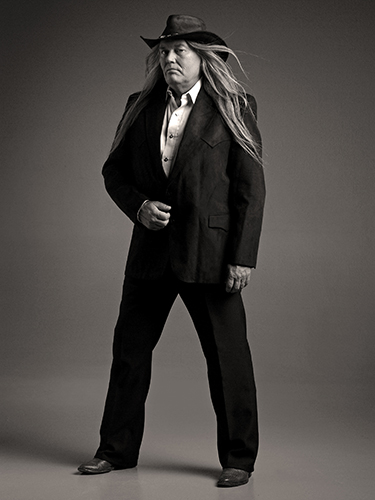 A man with long hair is wearing a wide-brimmed hat, a dark suit, and a light-colored shirt. He is standing proudly with his feet apart looking at the camera from the corner of his eyes.