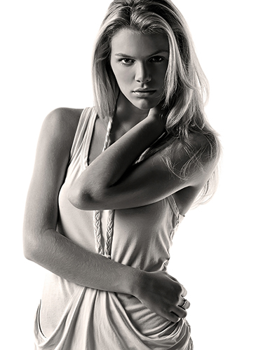 A woman with long hair, wearing a sleeveless top. She poses with one hand behind her neck and the other at her waist.