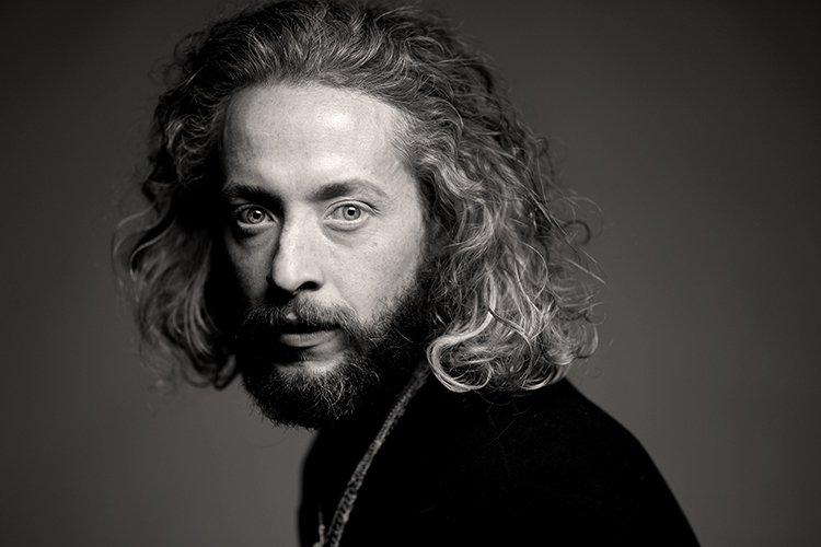 Portrait of a bearded man with long, curly hair looking towards the camera against a dark background. The shallow focus emphasizes his piercing eyes. 