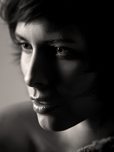 Close-up of a woman's face in soft focus. The lighting casts shadows across her features, emphasizing her eyes focused on something off-camera and her slightly parted lips.