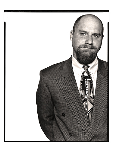 A black and white photo of a man with a beard, wearing a suit and an abstract-patterned tie. He is facing the camera with a slight smile, standing against a plain white background.