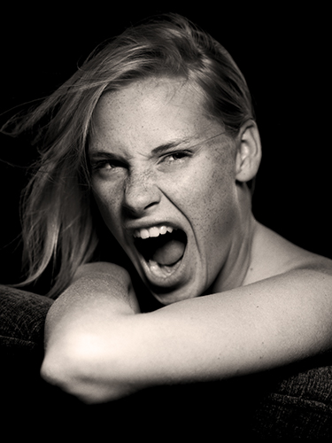 A black and white close-up of a person with light hair, expressing strong emotion with an open mouth and intense gaze. Their arm rests in front of them against a dark background.